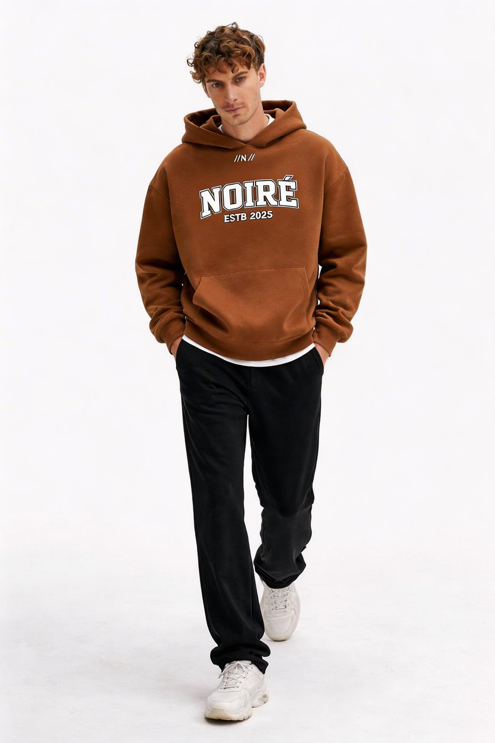 male model walking pose wearing oversized brown graphic streetwear hoodie noiré varsity logo design urban streetwear outfit