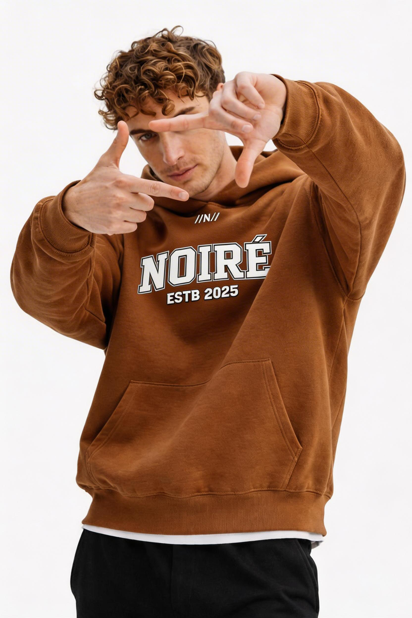 male model close up pose wearing oversized brown graphic streetwear hoodie noiré varsity logo streetwear fashion