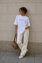 model leaning against wall wearing oversized white ballerina graphic t-shirt with pastel purple typography and cream pants streetwear fashion