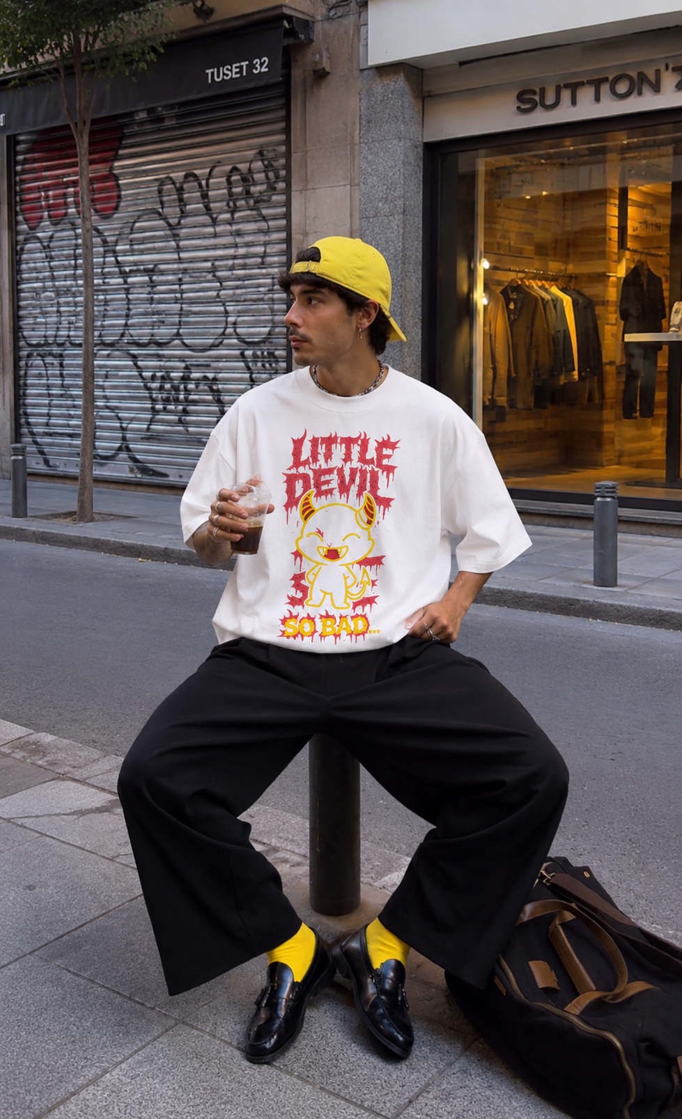 male model sitting on street bollard wearing oversized white graphic t shirt with little devil cartoon design and relaxed fit urban streetwear outfit