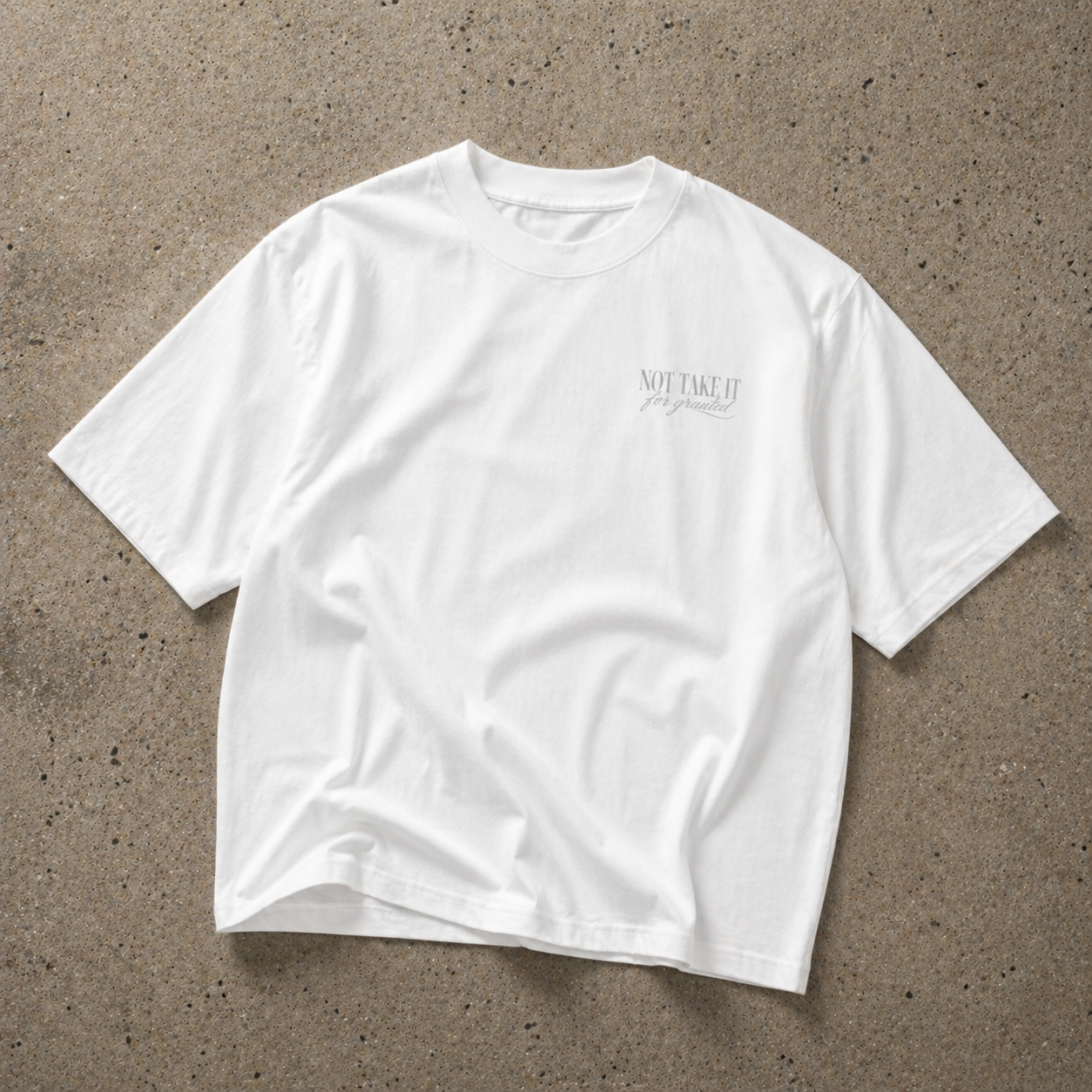front view oversized white t-shirt with minimalist grey “Not Take It For Granted” chest print laid flat on concrete background streetwear fashion