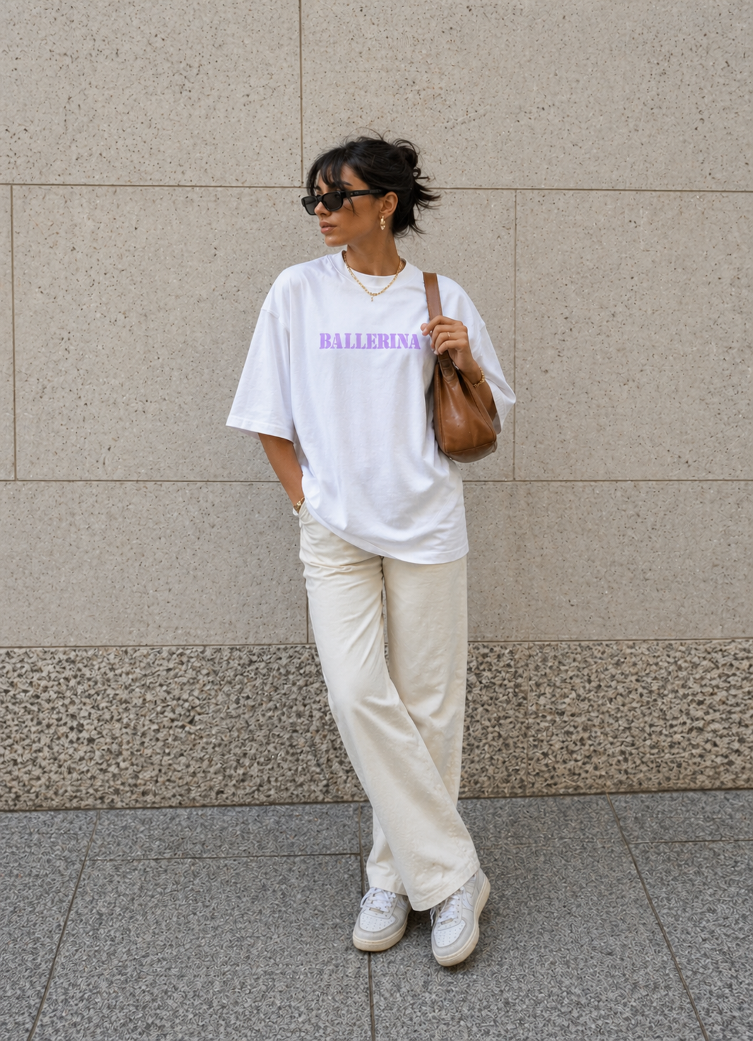model posing against wall wearing oversized white ballerina graphic t-shirt cream pants and brown shoulder bag urban streetwear outfit