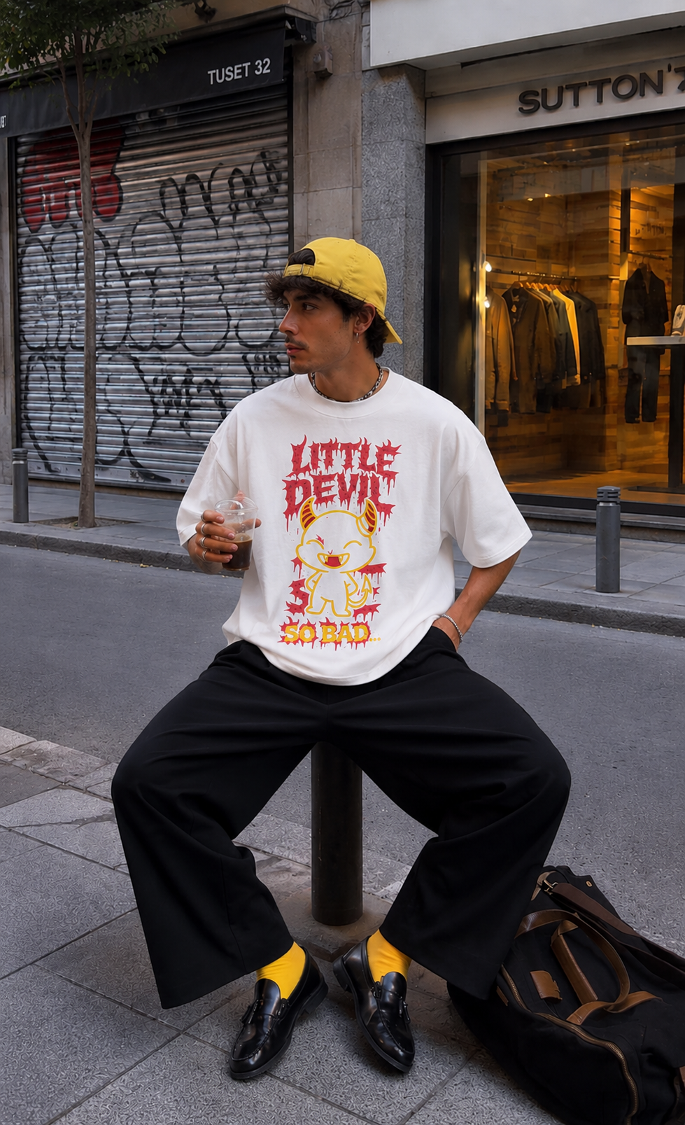 male model holding iced coffee wearing oversized white graphic t shirt featuring little devil graphic print streetwear fashion