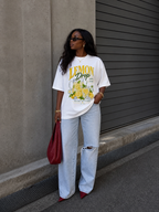 model standing side view wearing oversized white graphic t shirt lemon drop summer edition print with light wash ripped jeans streetwear fashion