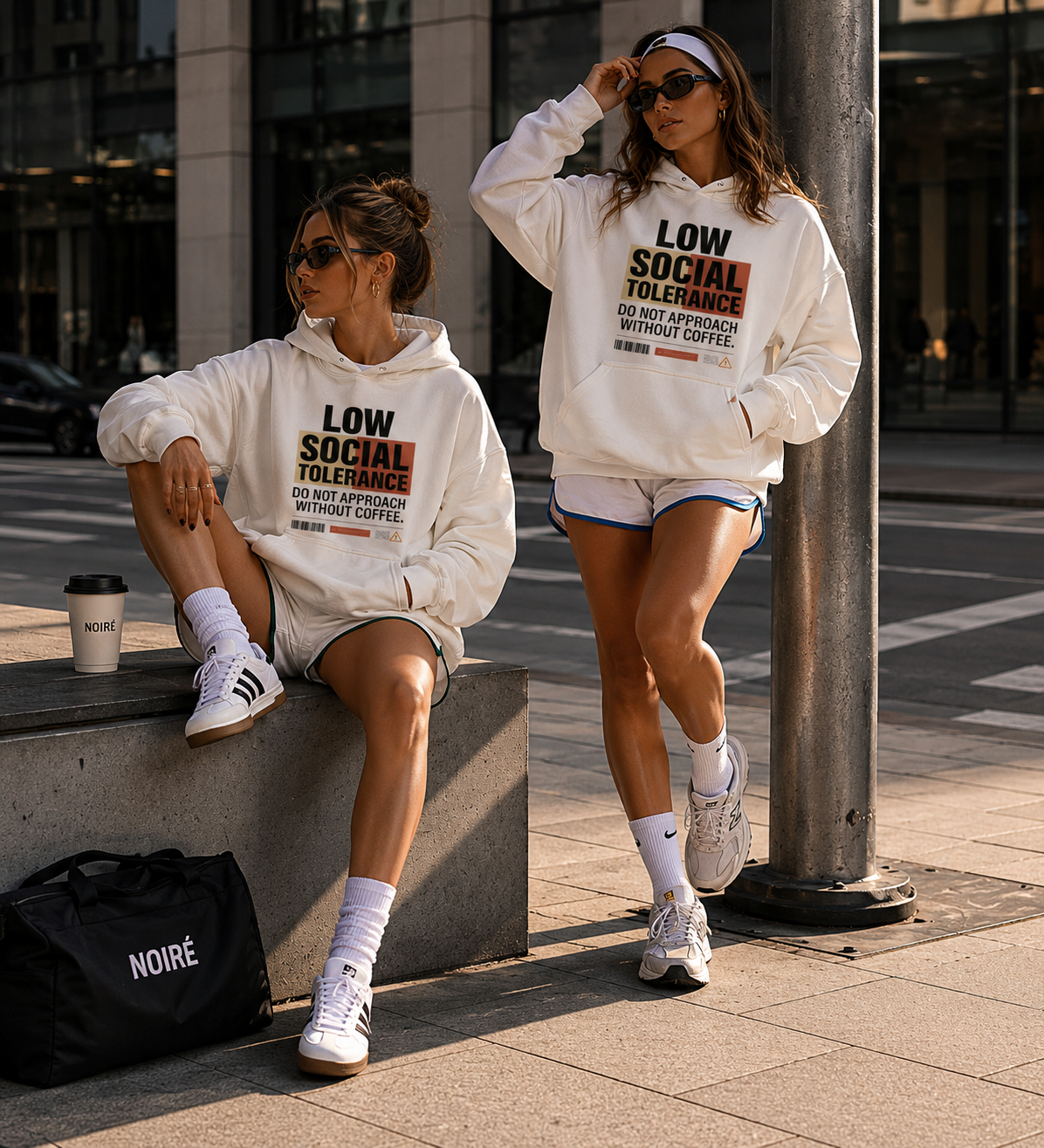 two women posing in oversized cream graphic hoodies with low social tolerance slogan modern streetwear fashion editorial