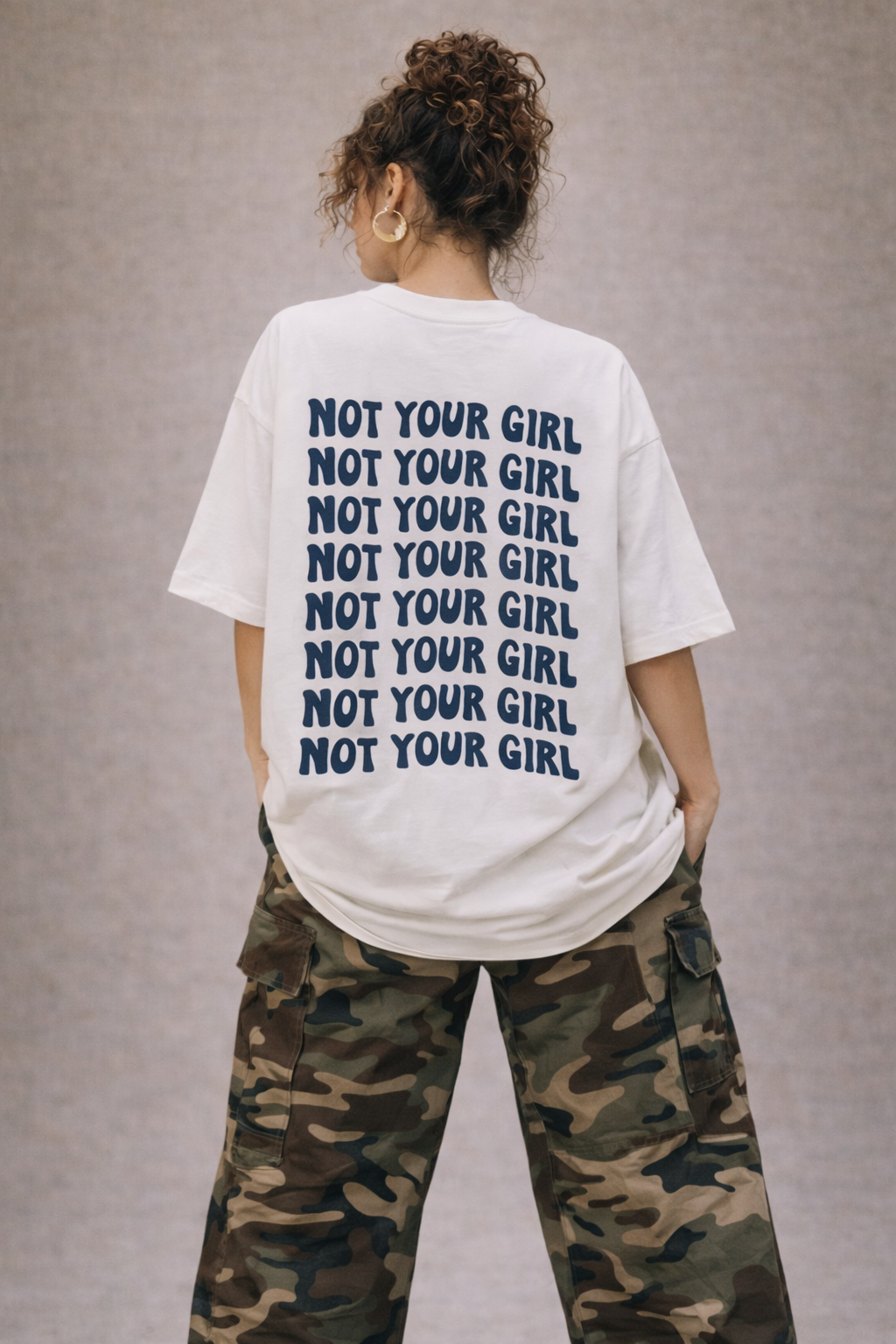 Back view of white oversized “Not Your Girl” statement t-shirt featuring stacked navy text design, styled with camouflage cargo pants, trendy women’s streetwear look.
