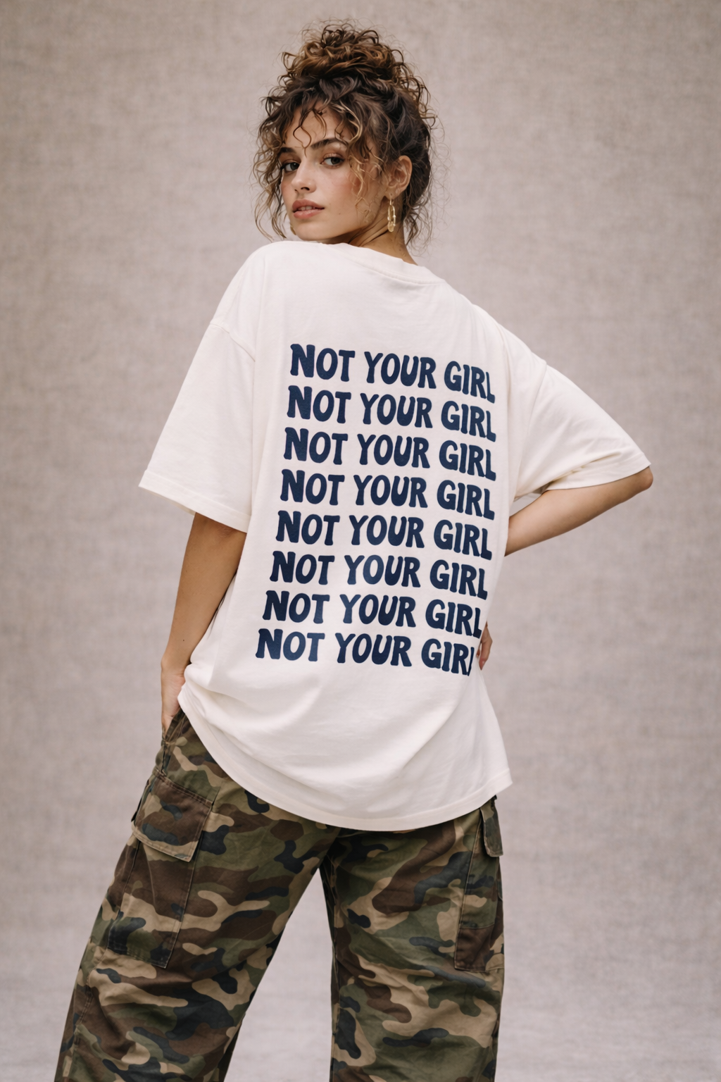 Woman wearing oversized white graphic t-shirt with repeated “Not Your Girl” navy typography on the back, styled with camouflage cargo pants, bold women’s streetwear outfit.