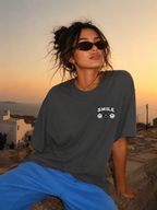 Woman leaning back wearing oversized charcoal graphic t-shirt front chest print, relaxed streetwear outfit overlooking the sea at sunset