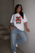 Woman leaning against wall wearing oversized white graphic t-shirt front view with Kiss Me Different Text Me Never graphic, oversized streetwear fashion