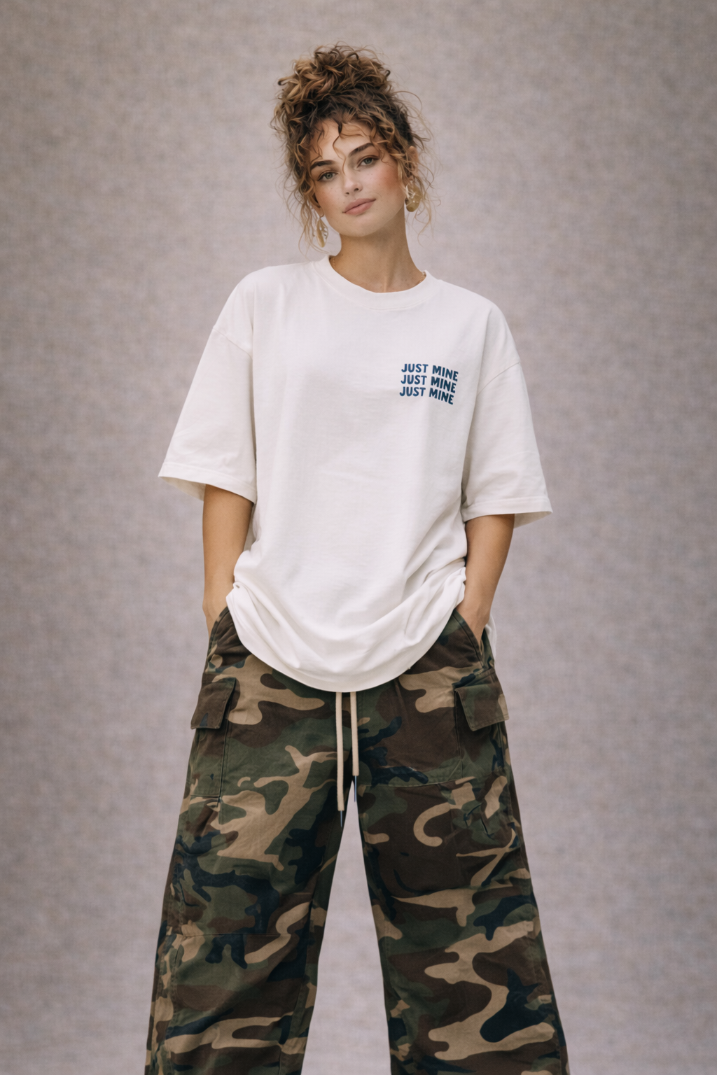 Female model in white oversized t-shirt with small “Just Mine” chest print, paired with camo cargo pants, modern women’s streetwear fashion.