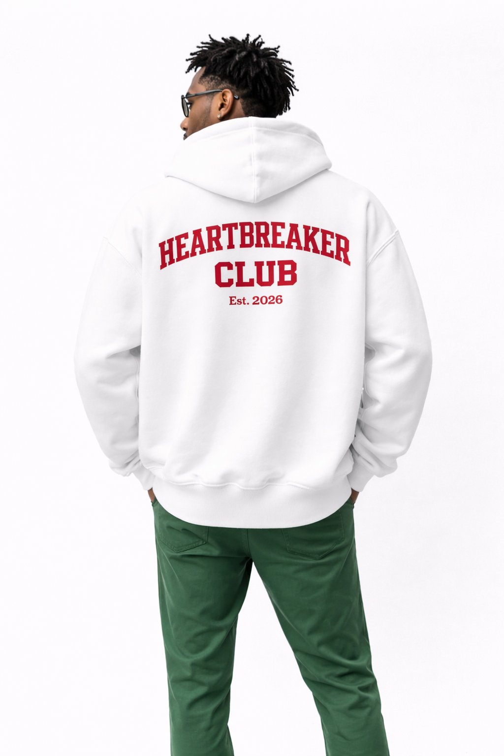 Man wearing white oversized hoodie with bold red “Heartbreaker Club” back print, modern streetwear outfit styled with green pants.