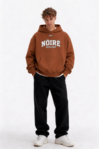 Male model wearing brown oversized Nöiré ESTB 2025 graphic hoodie, full-body front view, urban streetwear outfit