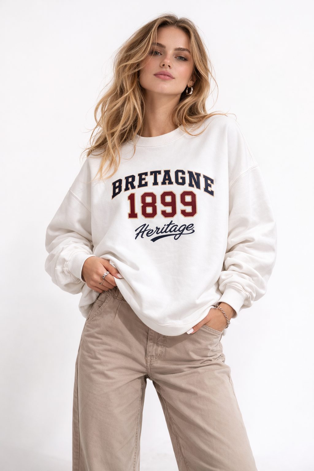 Woman wearing oversized cream sweatshirt with “Bretagne 1899 Heritage” varsity-style graphic print in navy and red, styled with beige trousers, casual heritage streetwear look.