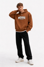Male model posing in brown oversized Nöiré collegiate logo hoodie, front view, graphic streetwear