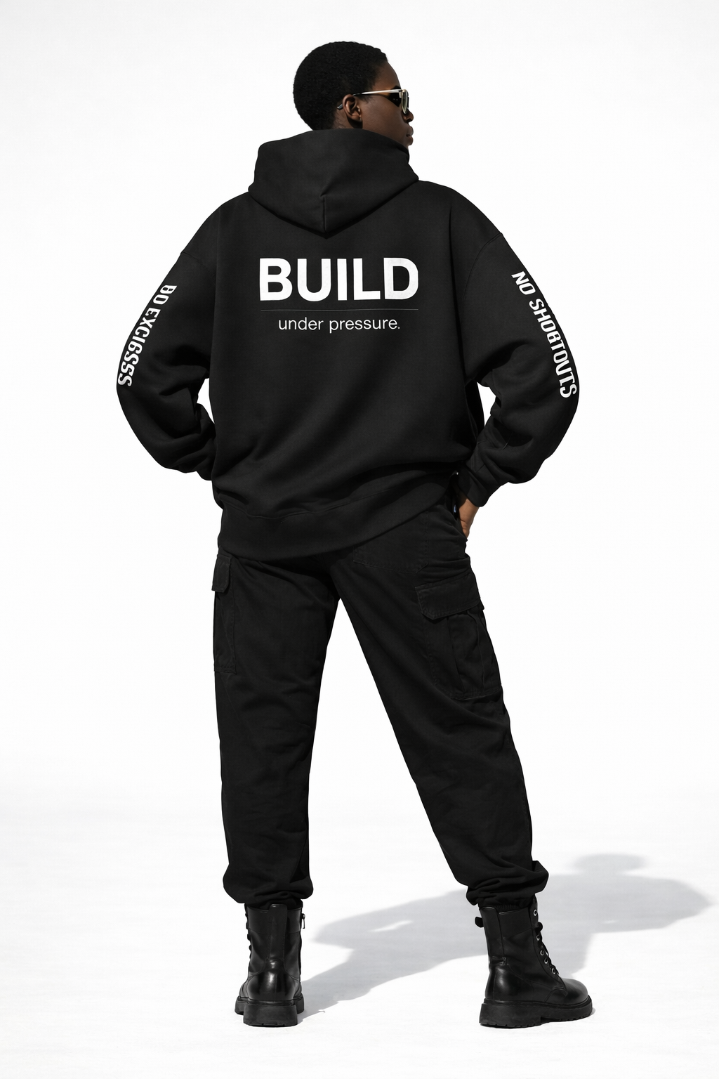 Back view of female model in black hoodie with “BUILD under pressure.” printed on back and “NO SHORTCUTS” sleeve detail, black cargo pants and boots, studio background.