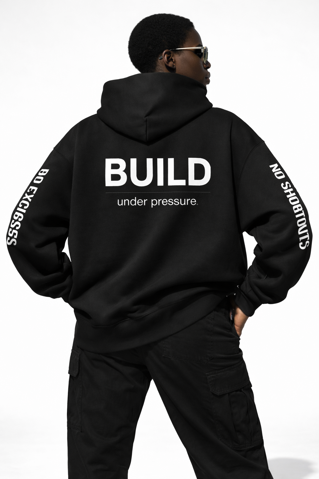 Close back view of female model wearing black hoodie with “BUILD under pressure.” back print and motivational sleeve text, studio lighting.