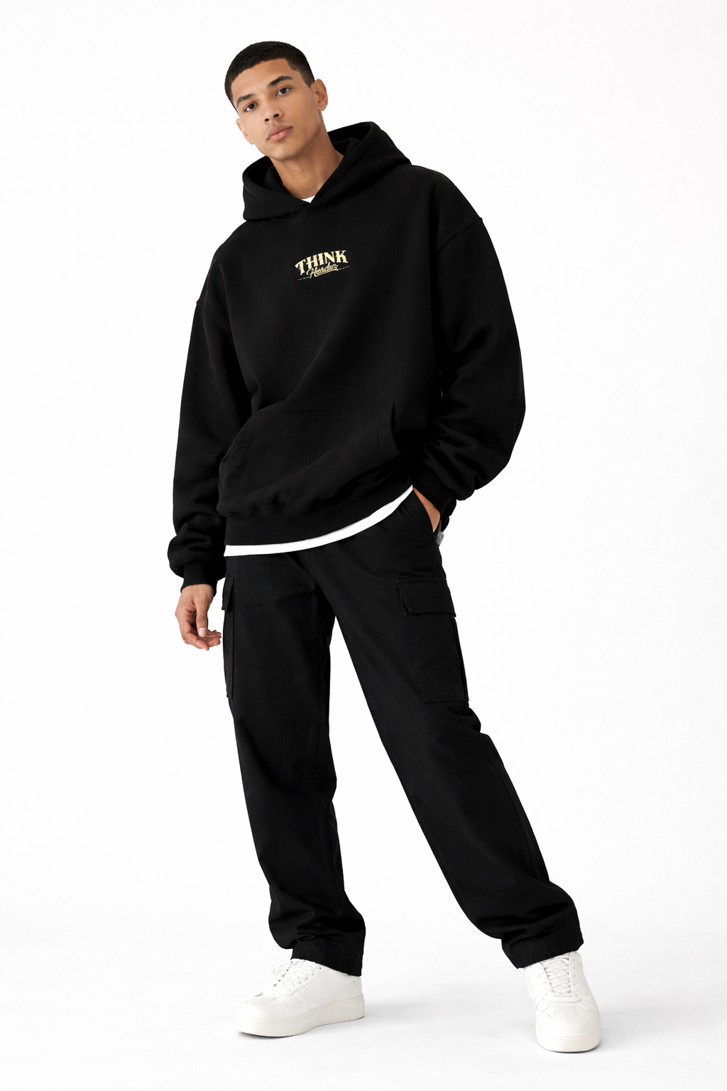 Male model wearing black oversized Noiré hoodie with small chest logo, full-body front view, black joggers and sneakers, minimal studio background.