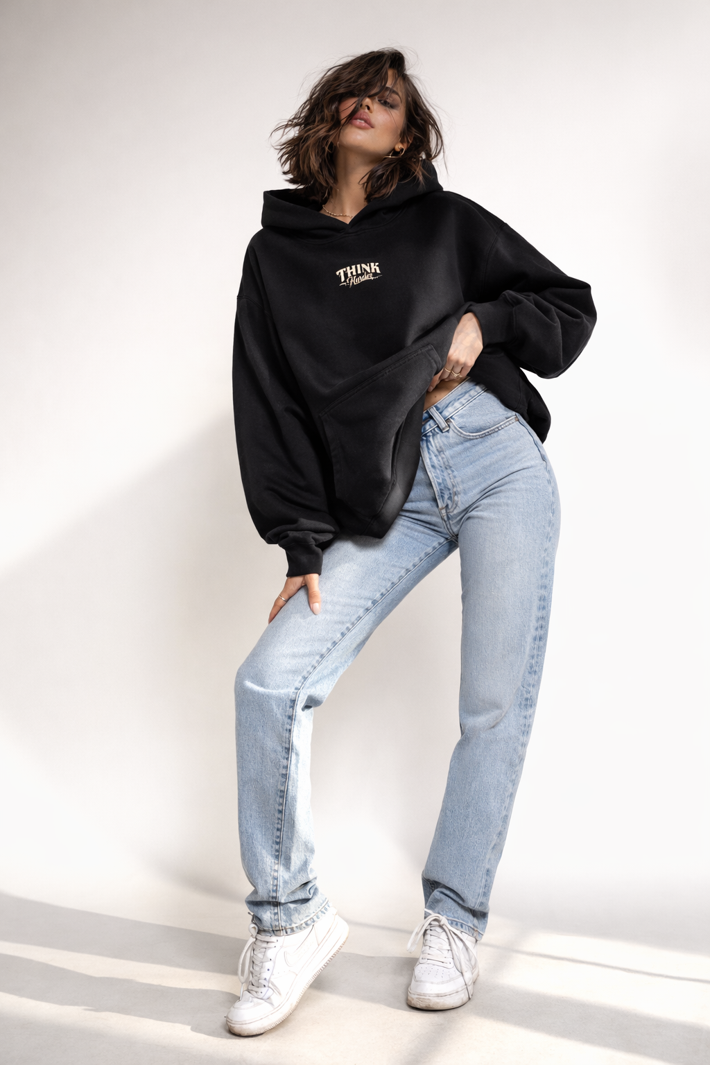 Female model wearing black oversized hoodie with small chest logo, casual streetwear styling with light blue jeans, studio background.