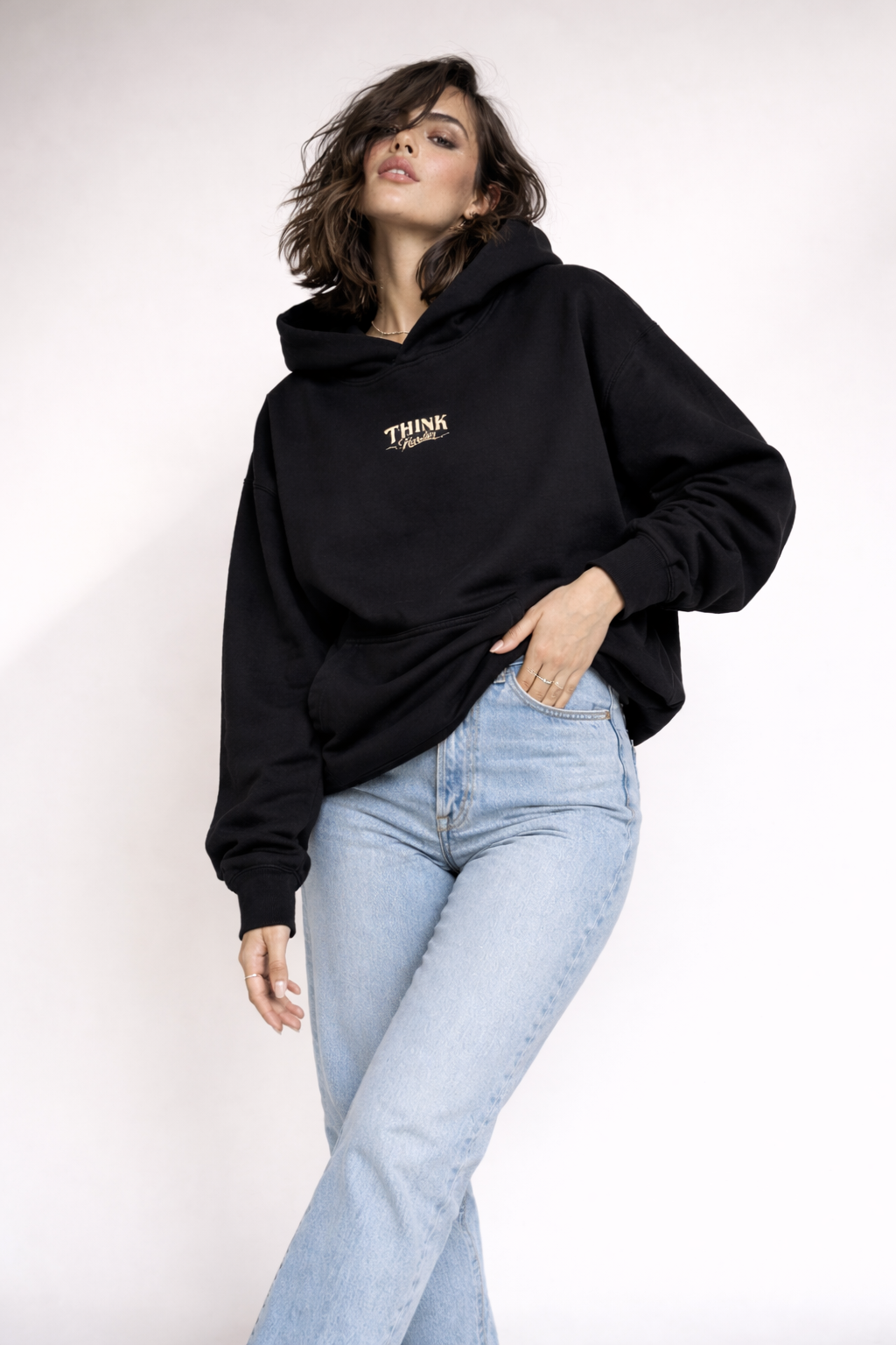 Female model posing in black oversized hoodie with subtle chest logo, casual denim styling, studio background.