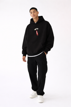 Black streetwear hoodie front view with small red and white chest graphic, male model wearing black cargo pants and white sneakers, modern urban fashion