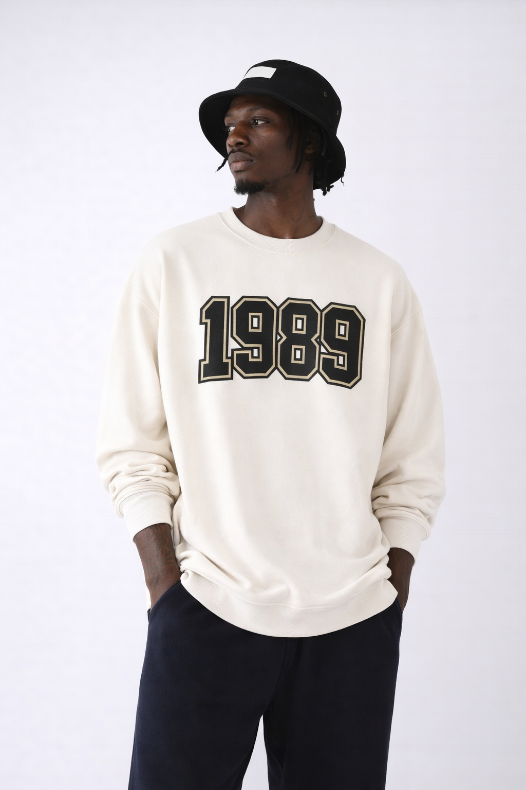 Man wearing beige oversized sweatshirt with 1989 varsity print, styled with black bucket hat and  pants, studio background – unisex streetwear sweatshirt 1989 design