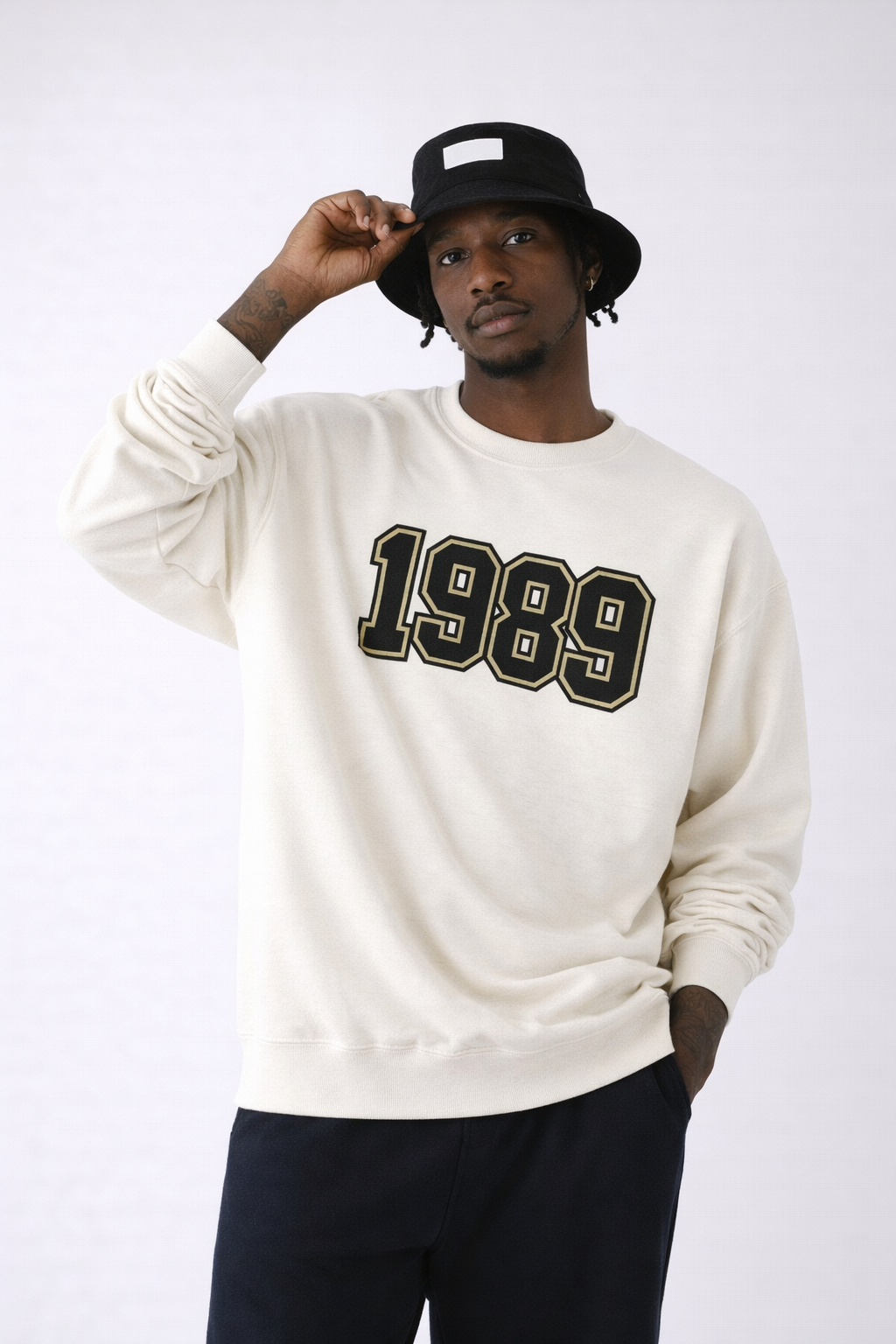 Male model in beige 1989 crewneck sweatshirt, relaxed fit with black and gold varsity numbers, minimalist studio fashion shoot