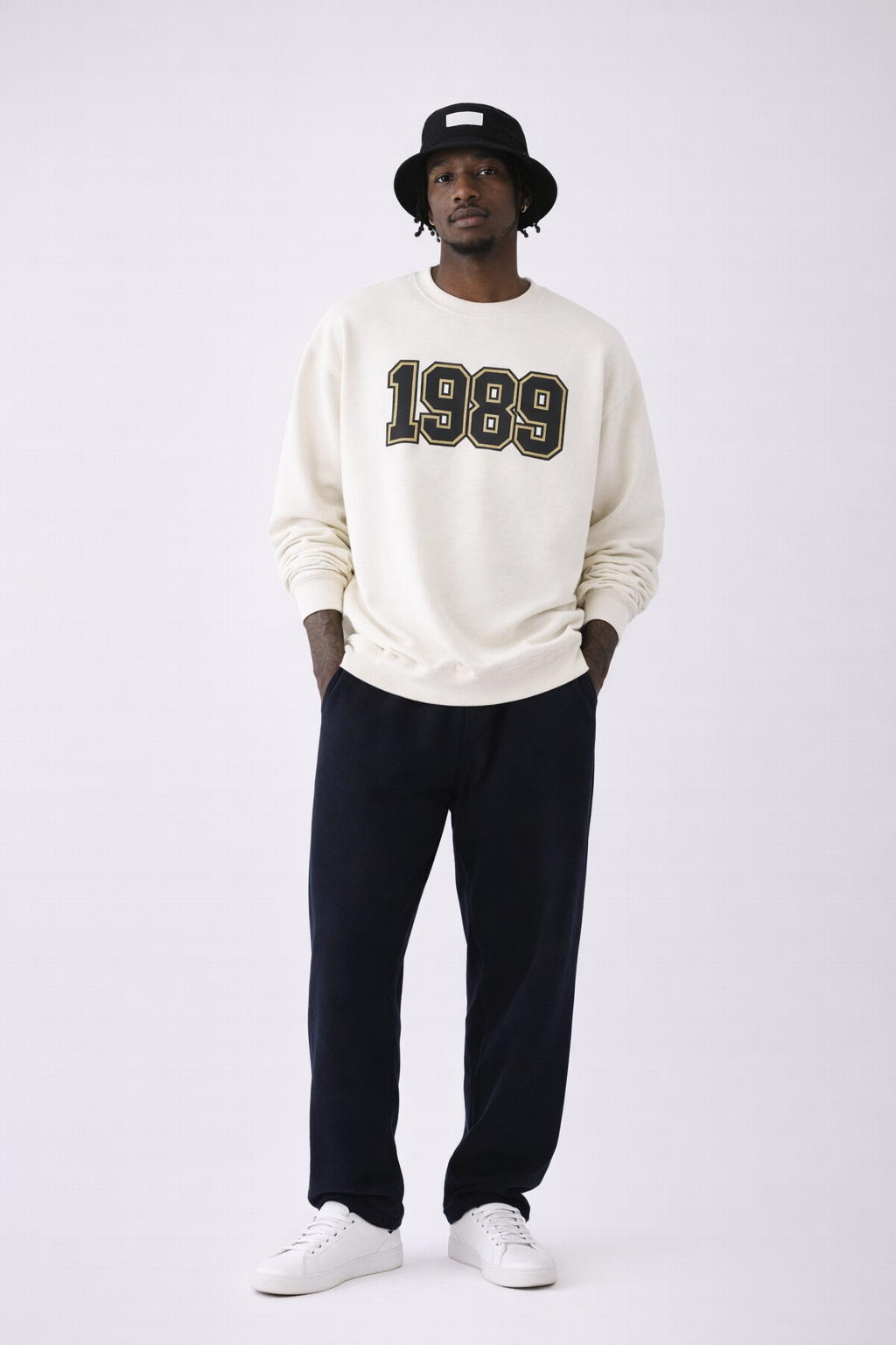 Full body shot of man in beige 1989 oversized sweatshirt paired with dark trousers and white sneakers, modern streetwear look
