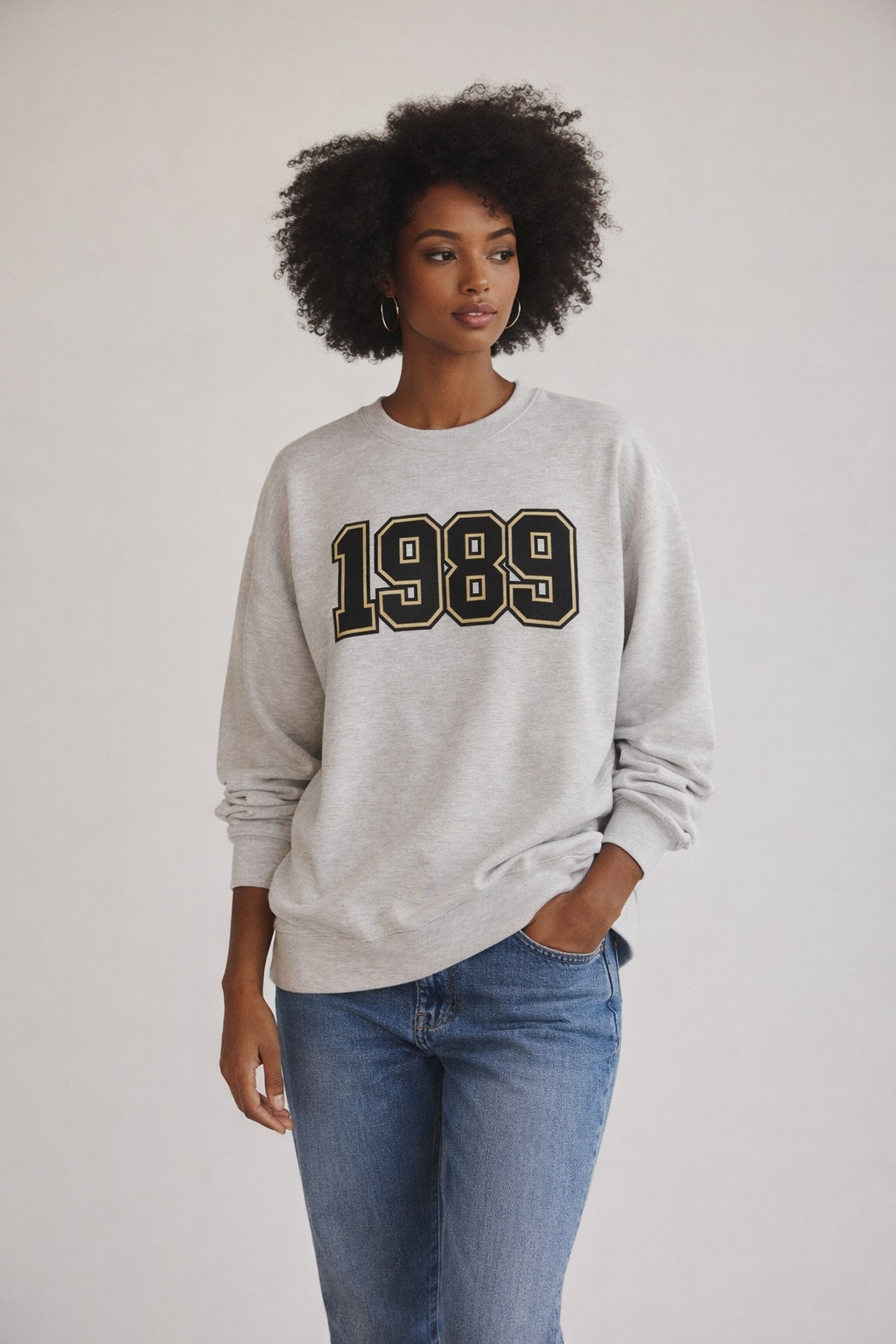 Woman wearing grey 1989 oversized sweatshirt with black and gold varsity print, styled with blue jeans, casual streetwear outfit