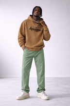 Men’s camel brown oversized hoodie with “Apocalypse” front graphic print, contemporary streetwear sweatshirt styled with green trousers, product image.