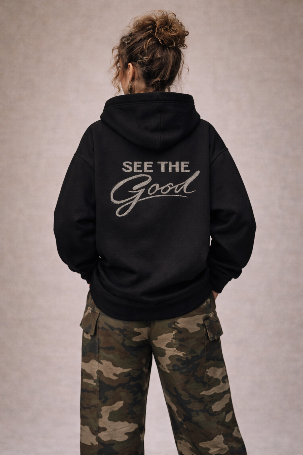 Female model in black oversized “See The Good” hoodie with large back graphic typography, paired with camo pants, modern women’s streetwear look.