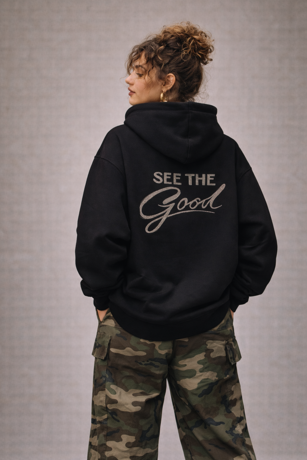 Woman wearing black oversized hoodie with “See The Good” back print in silver script, styled with camouflage cargo pants, women’s streetwear outfit rear view.