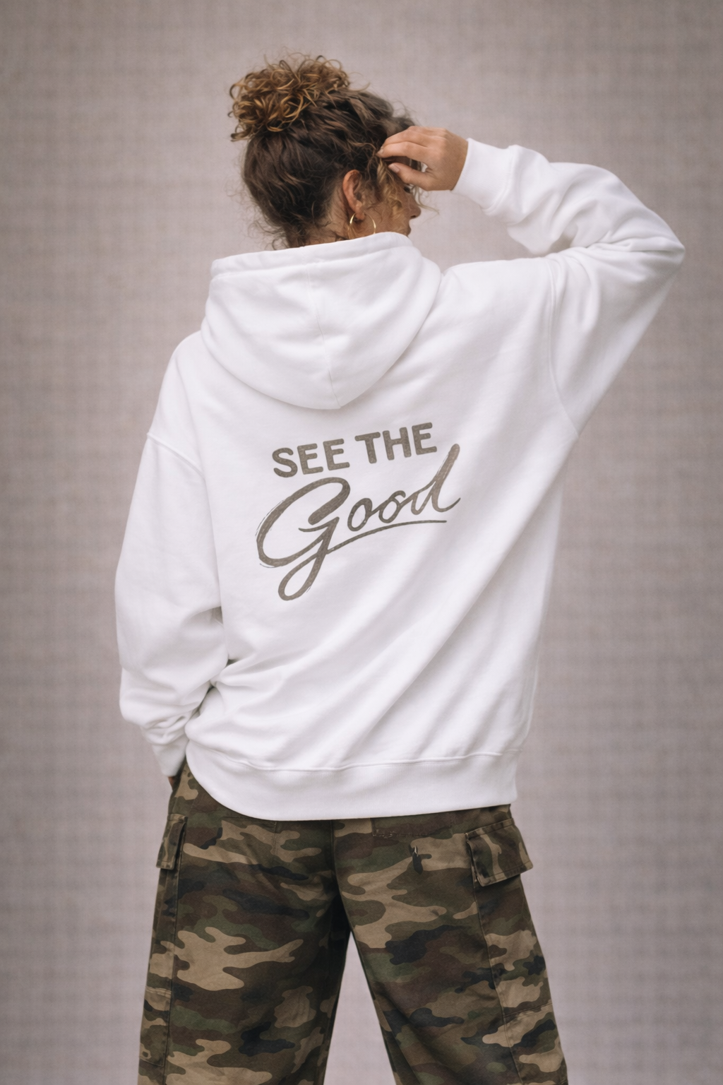 Woman wearing white oversized hoodie with “See The Good” statement back print in neutral tone, styled with camouflage cargo pants, women’s streetwear fashion.