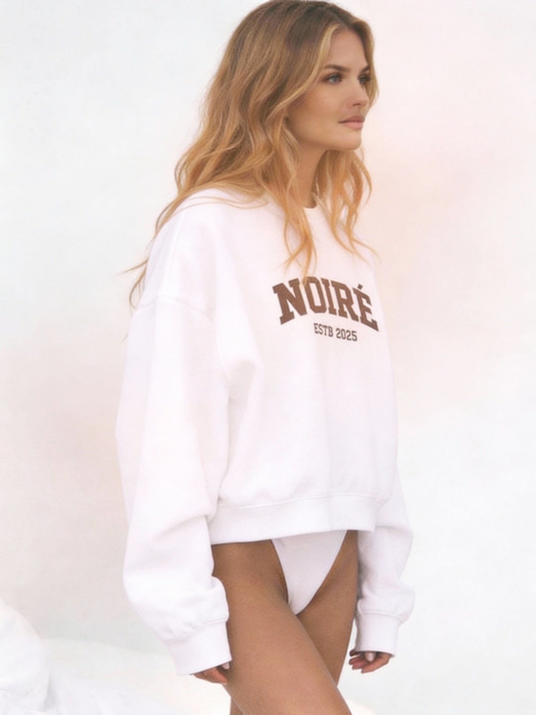 Female model walking while wearing white Noiré oversized sweatshirt, minimalist studio background, casual urban outfit.