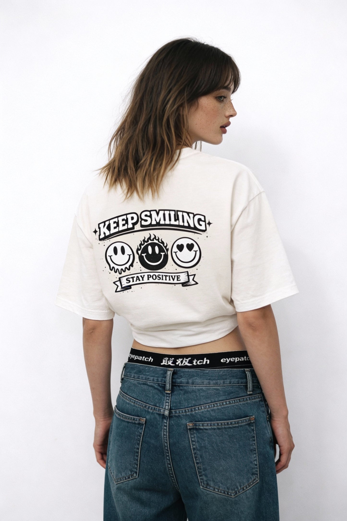 Model wearing beige t-shirt showing “Keep Smiling – Stay Positive” back print with smiley faces, creative streetwear look by Noiré.