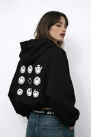 Woman wearing black hoodie with smiley grid graphic on the back, casual streetwear outfit photographed in studio.