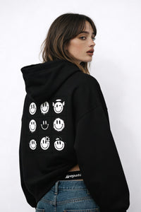 Woman wearing black hoodie with smiley grid graphic on the back, casual streetwear outfit photographed in studio.