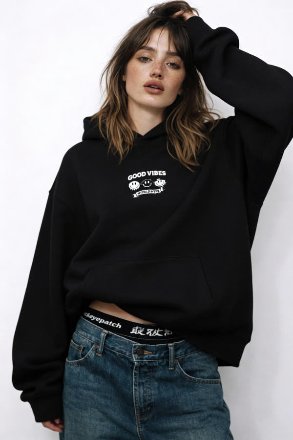 Woman wearing black hoodie with small smiley logo on chest, modern casual streetwear look in studio setting.