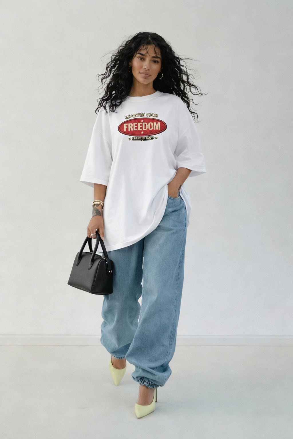 Woman wearing oversized white graphic T-shirt with “Imported From Freedom – Premium Goods” vintage logo on the front, styled with loose blue jeans and heels, casual streetwear outfit.