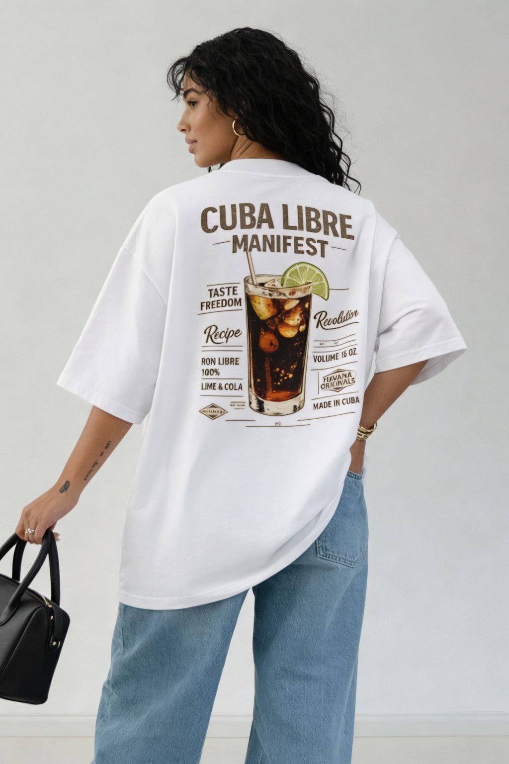 Back view of woman wearing oversized white T-shirt with large “Cuba Libre Manifest” cocktail graphic print, relaxed fit streetwear look with wide-leg jeans.
