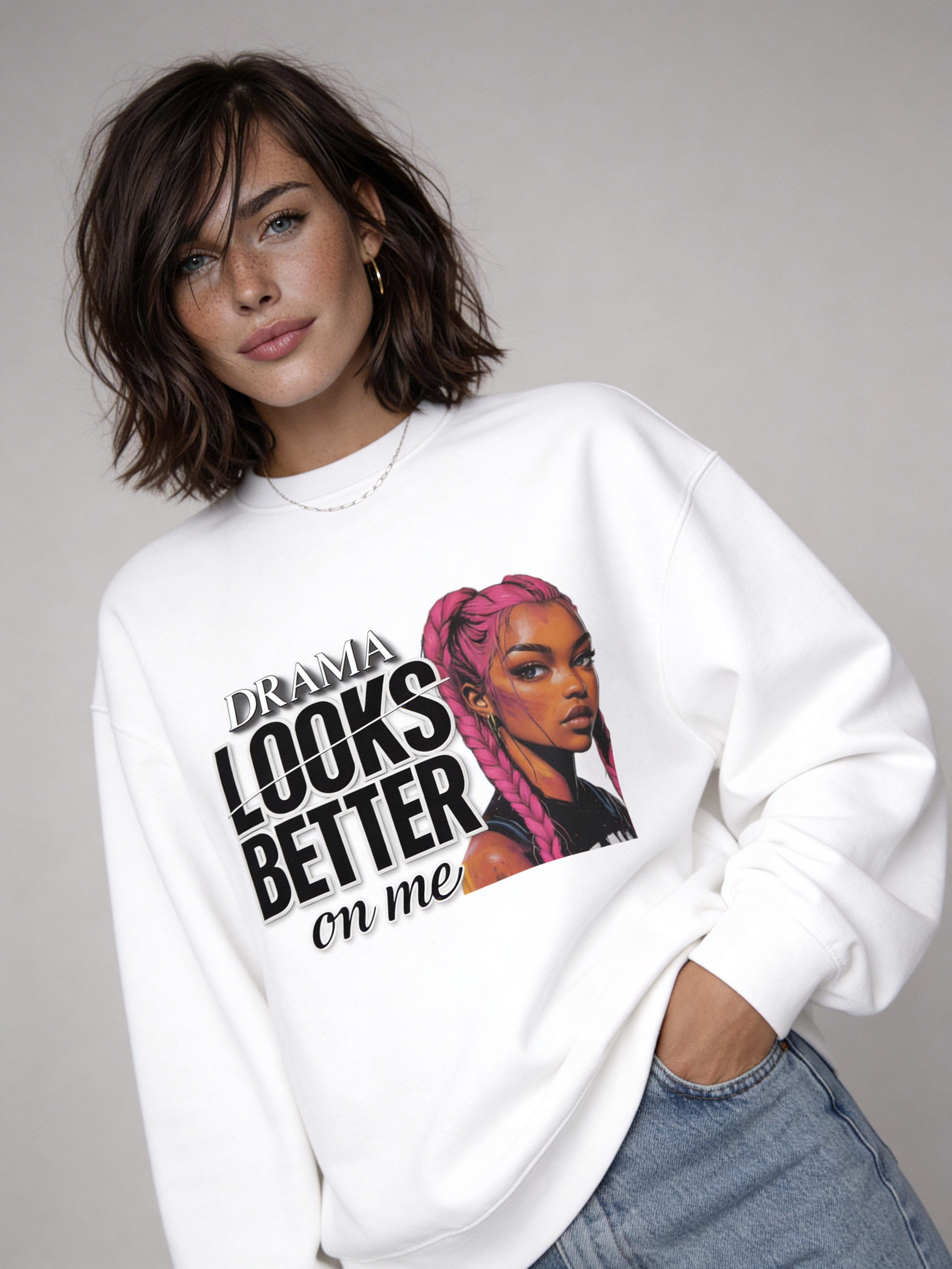 White oversized sweatshirt with “DRAMA LOOKS BETTER ON ME” graphic and illustrated girl with pink braids, worn by female model with light blue jeans, studio portrait.