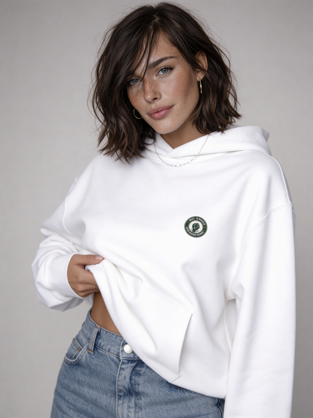 White oversized hoodie with small green Noiré emblem on chest, front view on female model, streetwear sweatshirt