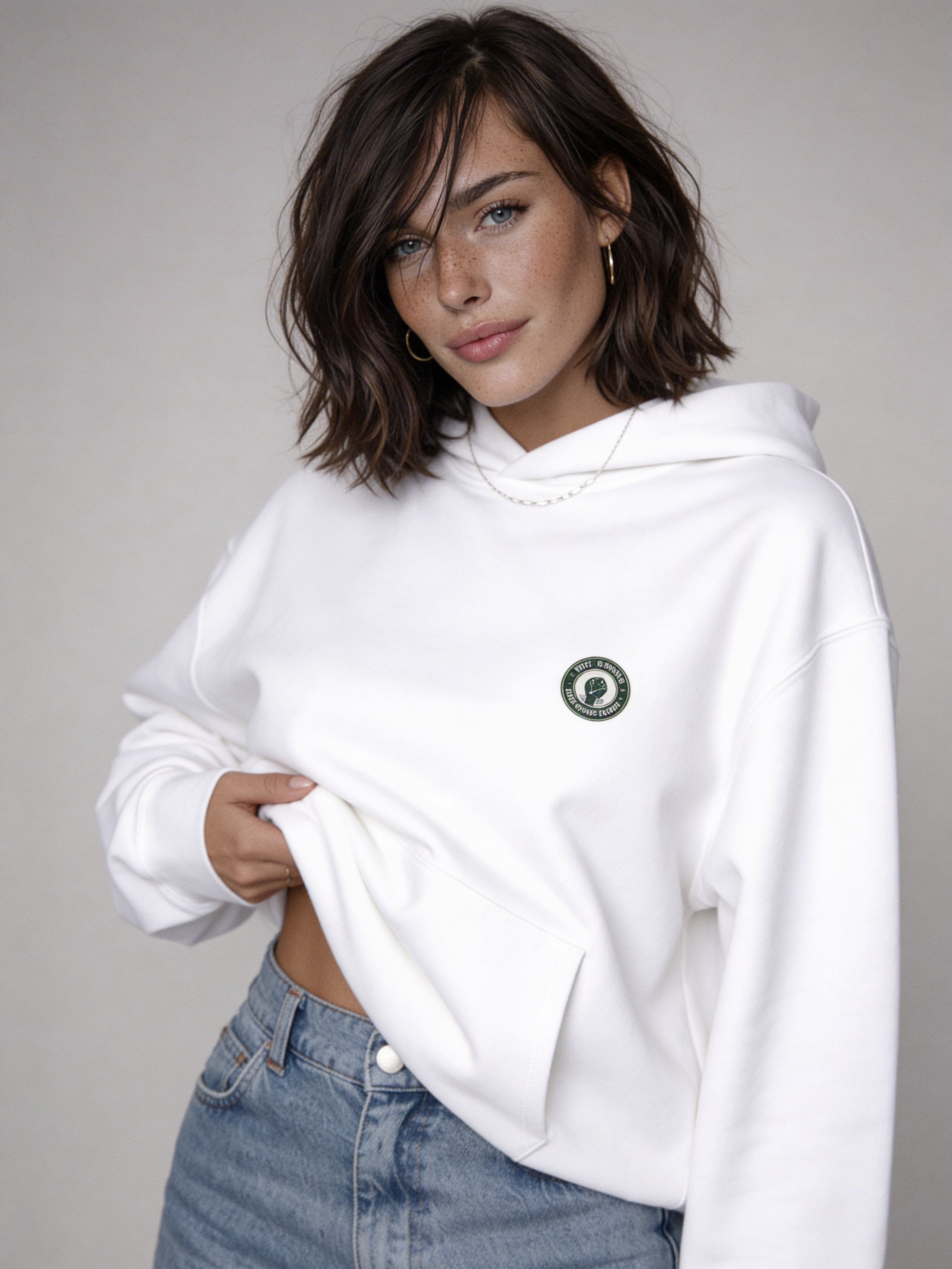 White oversized hoodie with small green Noiré emblem on chest, front view on female model, streetwear sweatshirt