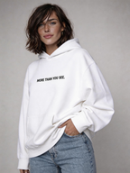 Woman wearing oversized white hoodie with “More Than You See” front text print, styled with light wash jeans, minimalist women’s streetwear outfit product photo.
