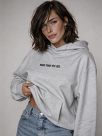 Female model wearing oversized light grey hoodie with “More Than You See” front text print, styled with light blue denim jeans, women’s streetwear fashion product image for online store.