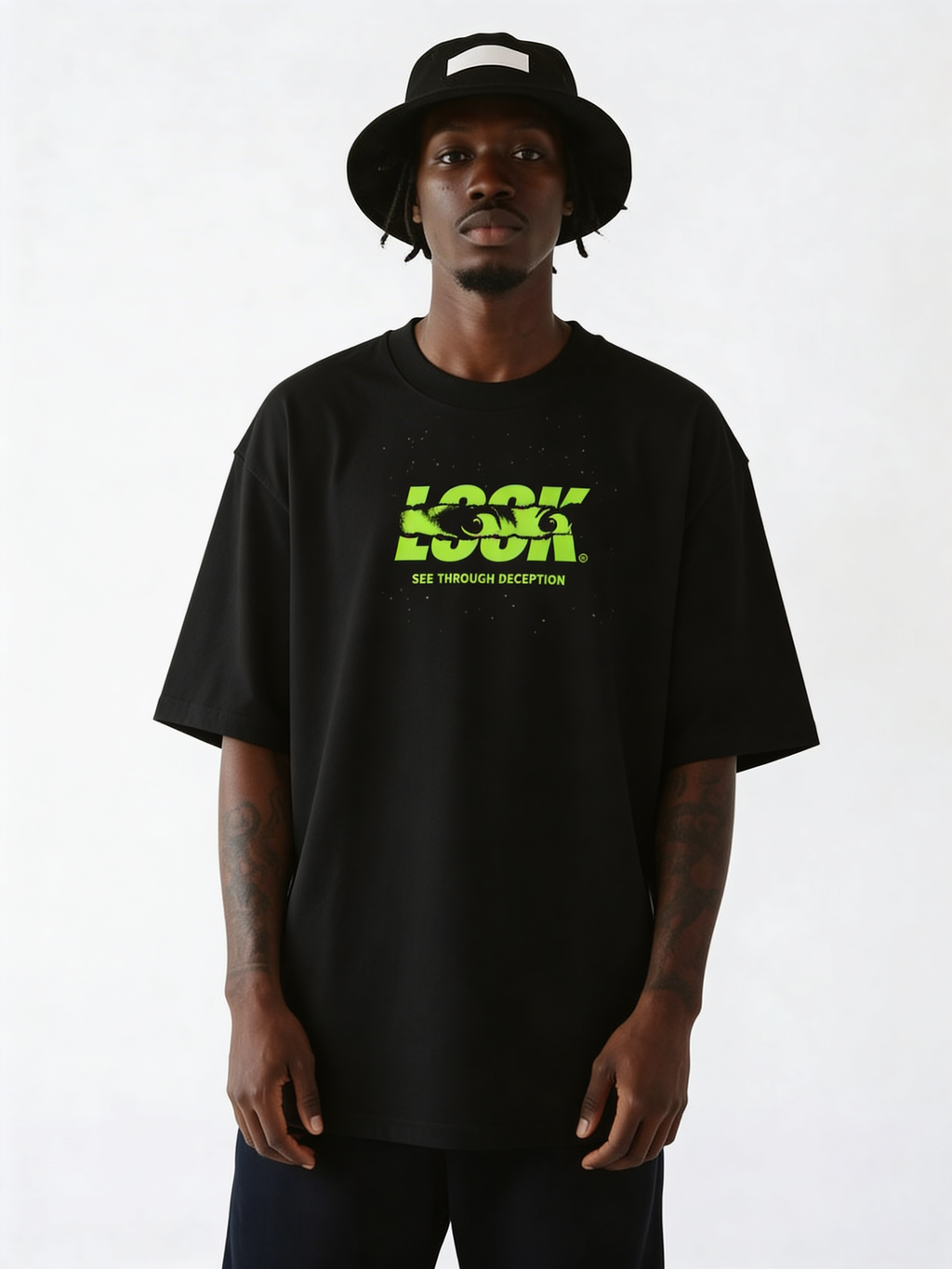 Black oversized streetwear T-shirt with neon green “LOOK – See Through Deception” graphic print on front, worn by male model with bucket hat, urban minimalist studio background.