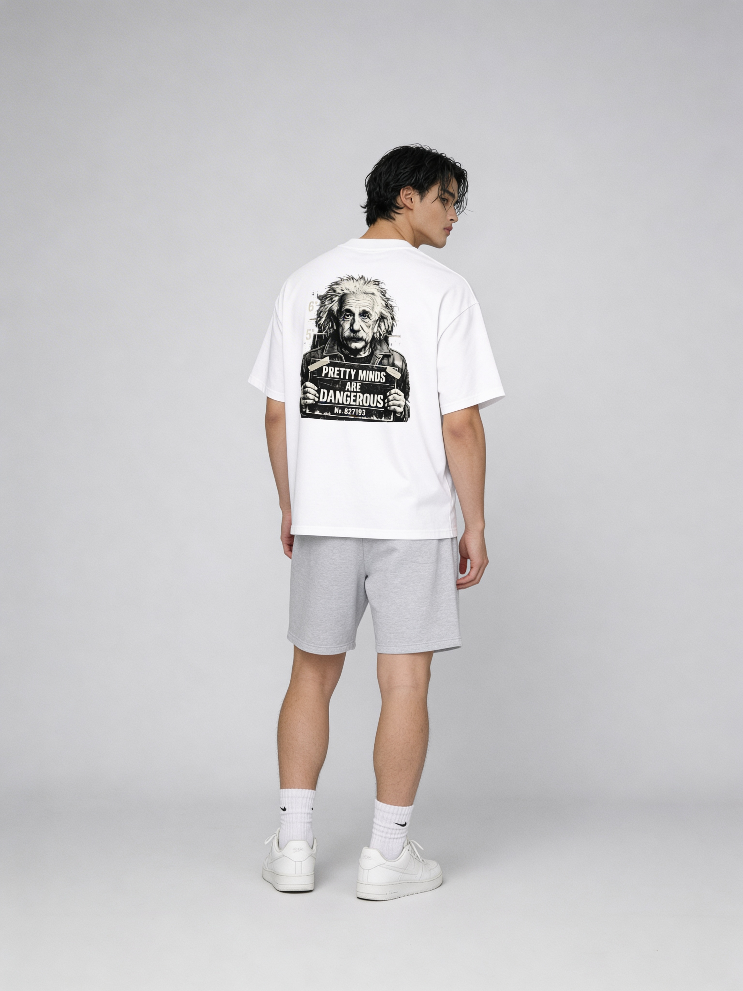 Male model wearing oversized white streetwear t-shirt with Albert Einstein mugshot graphic and “Pretty Minds Are Dangerous” back print, styled with grey shorts and white sneakers in studio