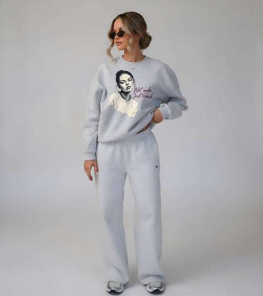 Female model wearing grey oversized sweatshirt with illustrated female face and "Not nude, just French" text, studio lifestyle shot