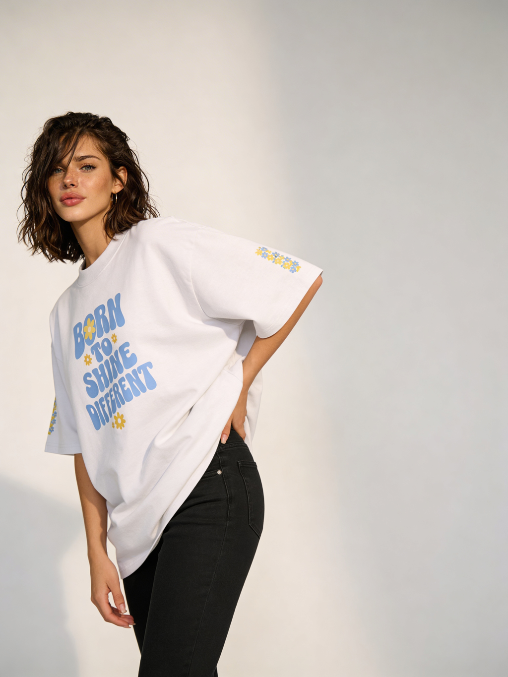 Female model wearing oversized white “Born to Shine Different” graphic t-shirt with floral accents on sleeves, styled with black jeans in studio