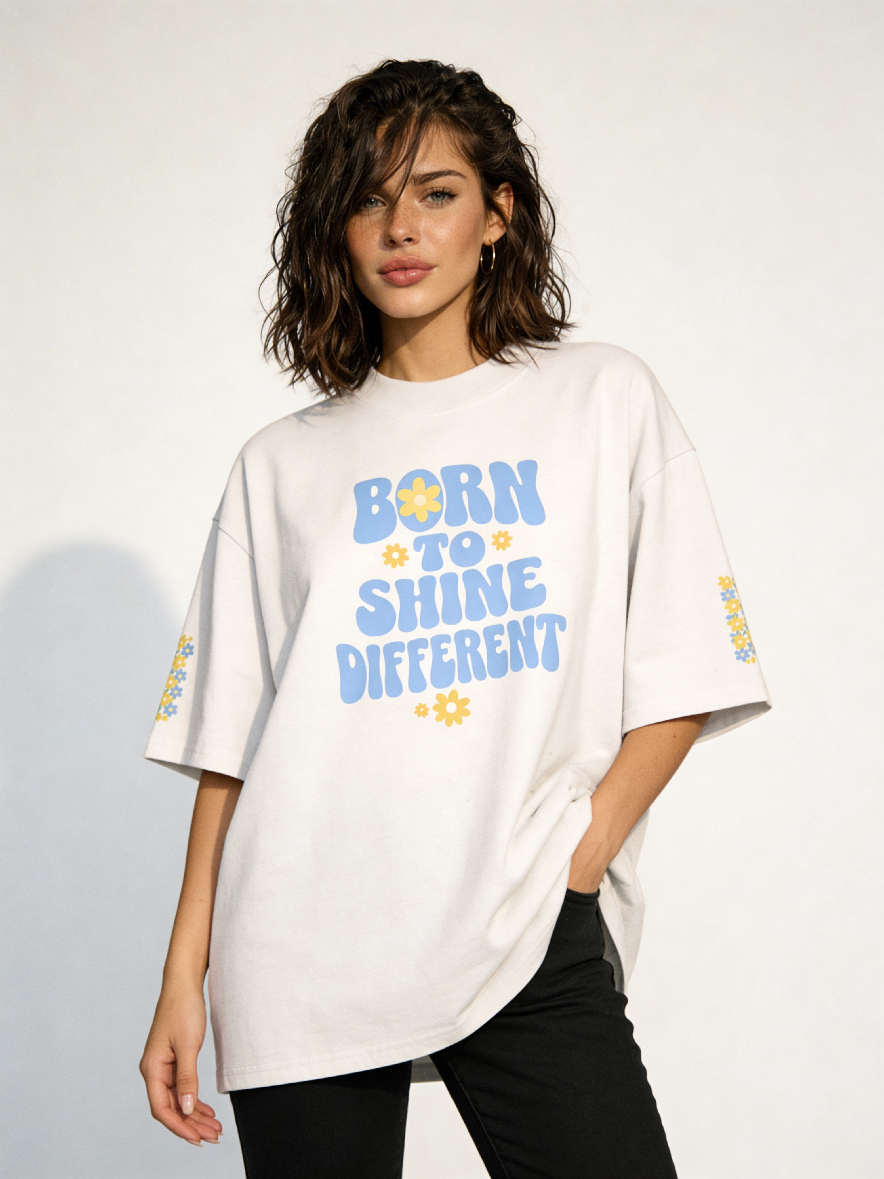 Front view of woman in oversized white motivational quote tee featuring blue and yellow “Born to Shine Different” print, modern casual outfit