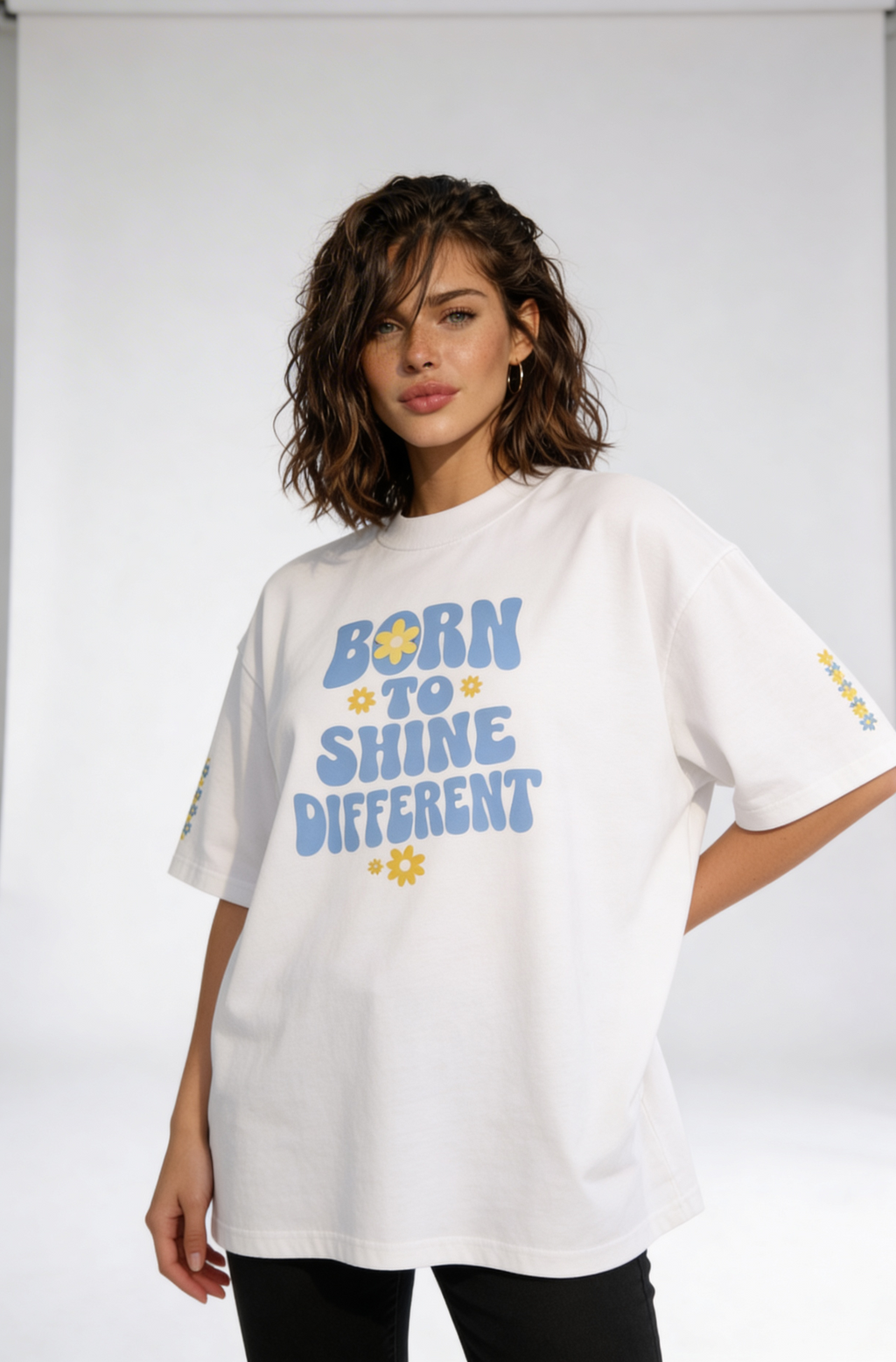 Female model in oversized white streetwear t-shirt with “Born to Shine Different” pastel typography and daisy graphics, contemporary women’s fashion